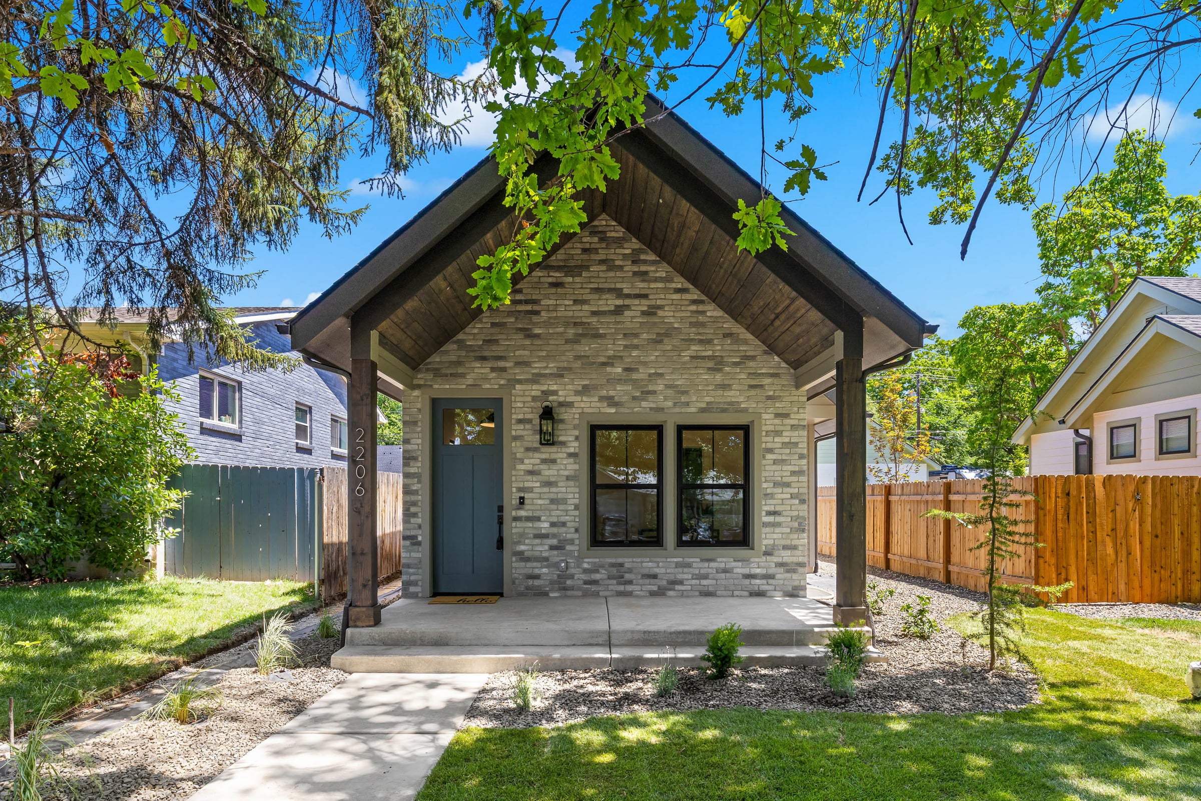 Street view of the 20th Street Residence in Boise: a brick cottage with a deep gabled porch on timber posts, a blue-green front door, and mature trees overhead.