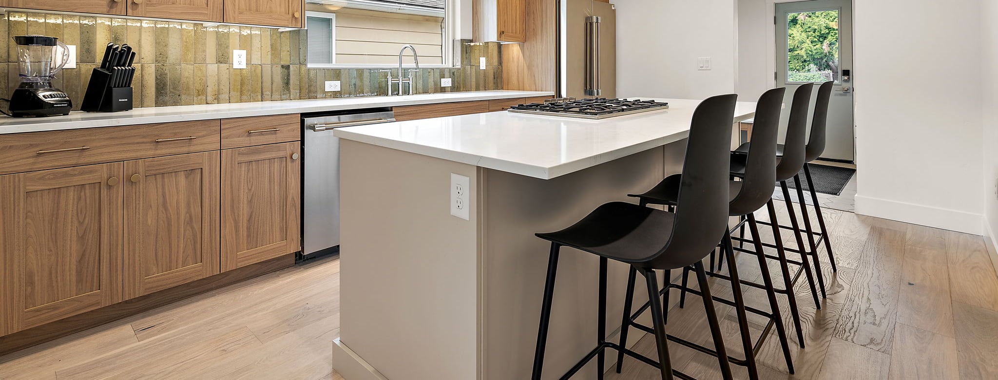 Counter-height view across the 25th Street ADU kitchen: the white quartz island with four black stools, walnut lower cabinets, and white oak flooring.