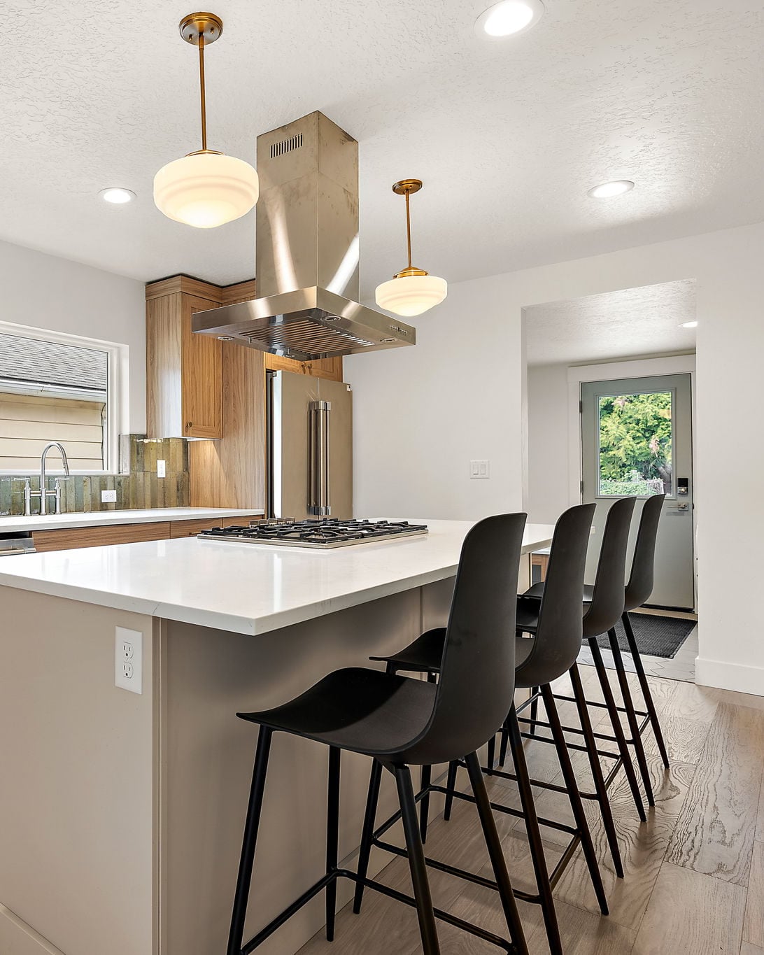 Kitchen of the 25th Street ADU in Boise: a white quartz island with black stools, a stainless island hood, and schoolhouse pendants, looking toward the back door.