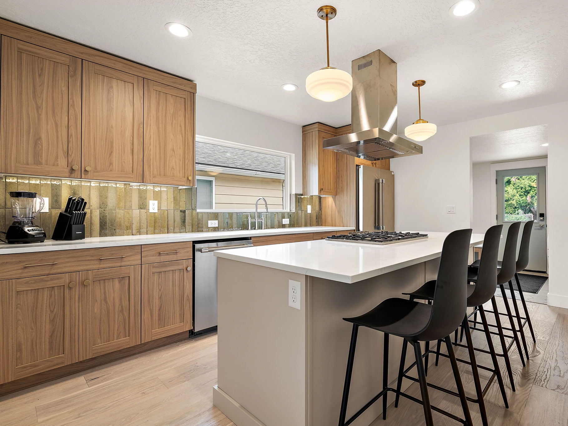 Walnut cabinetry and a green glazed tile backsplash in the 25th Street ADU kitchen, with the island and stainless hood to the right.