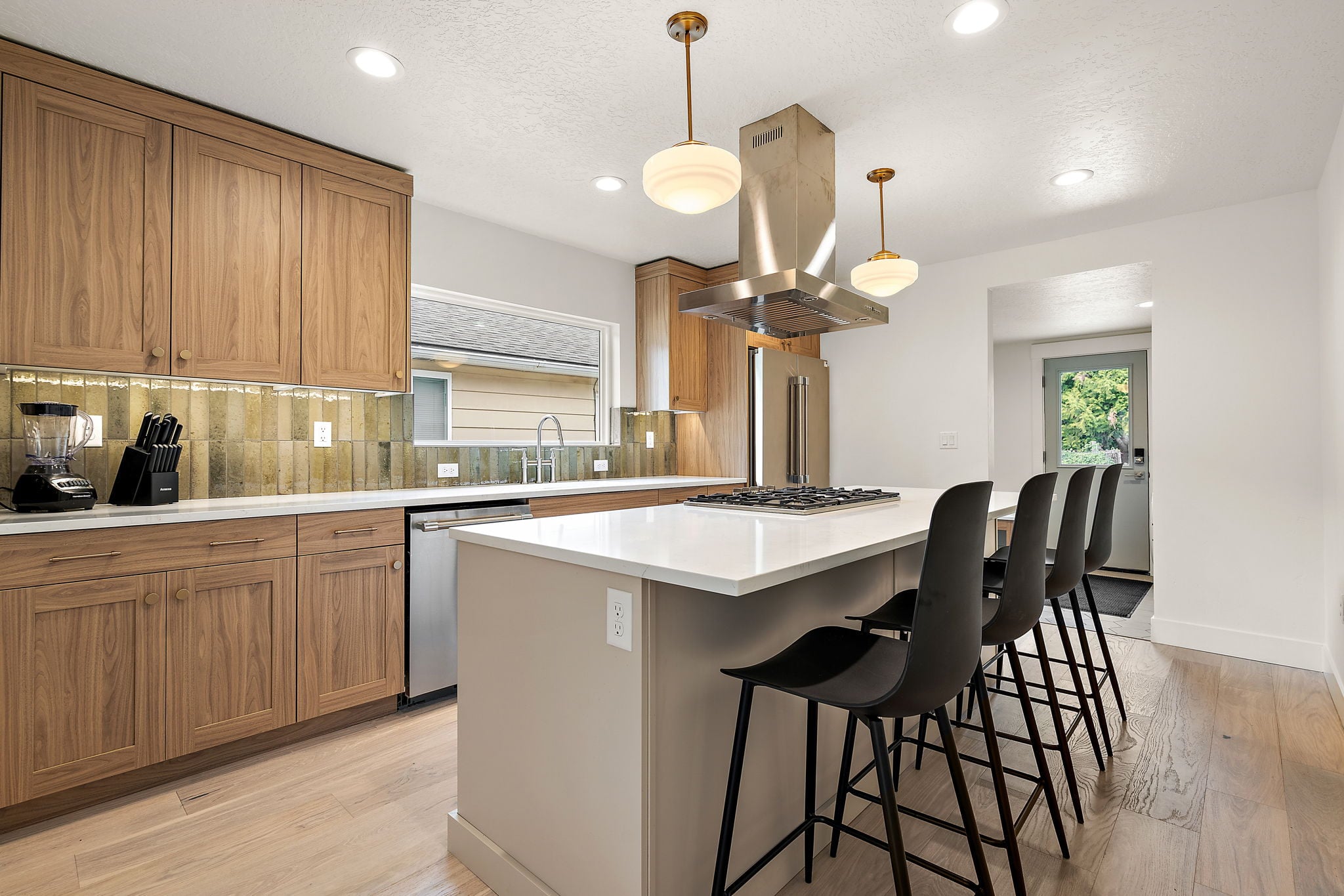 Kitchen of the 25th Street ADU in Boise: walnut cabinetry and a green glazed tile backsplash on the left, a white quartz island with black stools and a stainless island hood, and the back door beyond.